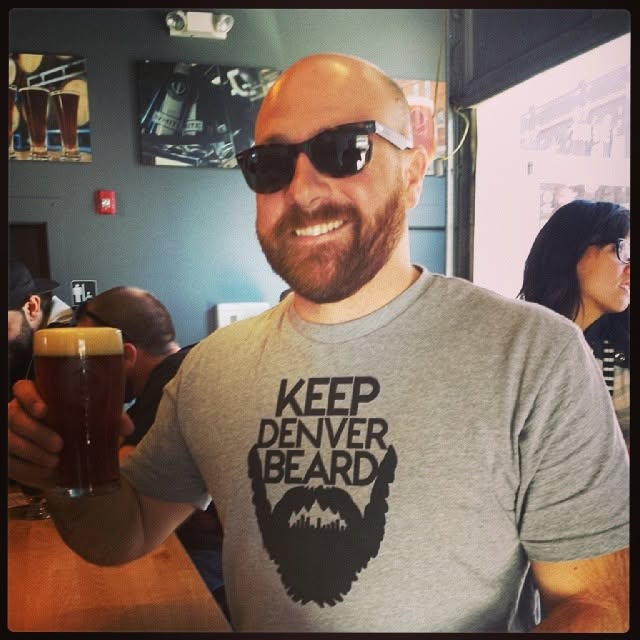 A man in sunglasses holding a beer while wearing a gray Keep Denver Beard T-shirt