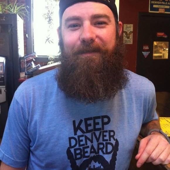 A man with a full beard wearing a blue Keep Denver Beard T-shirt inside a Denver bar