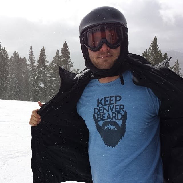 A skier opening his jacket to reveal a Keep Denver Beard T-shirt on the slopes